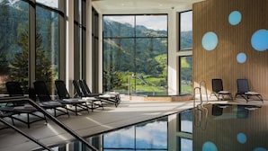 A modern indoor pool area with clear glass walls, lounge chairs, and a view of a mountainous landscape. - Hotel Goldried (Matrei in Osttirol)