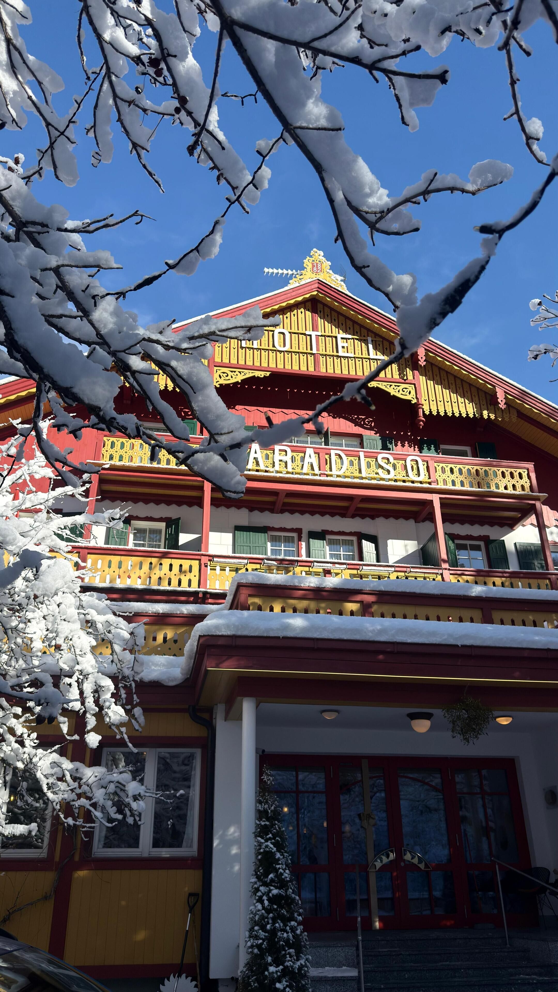 A hotel with a red and yellow facade, balconies, and snow-covered branches in the foreground.