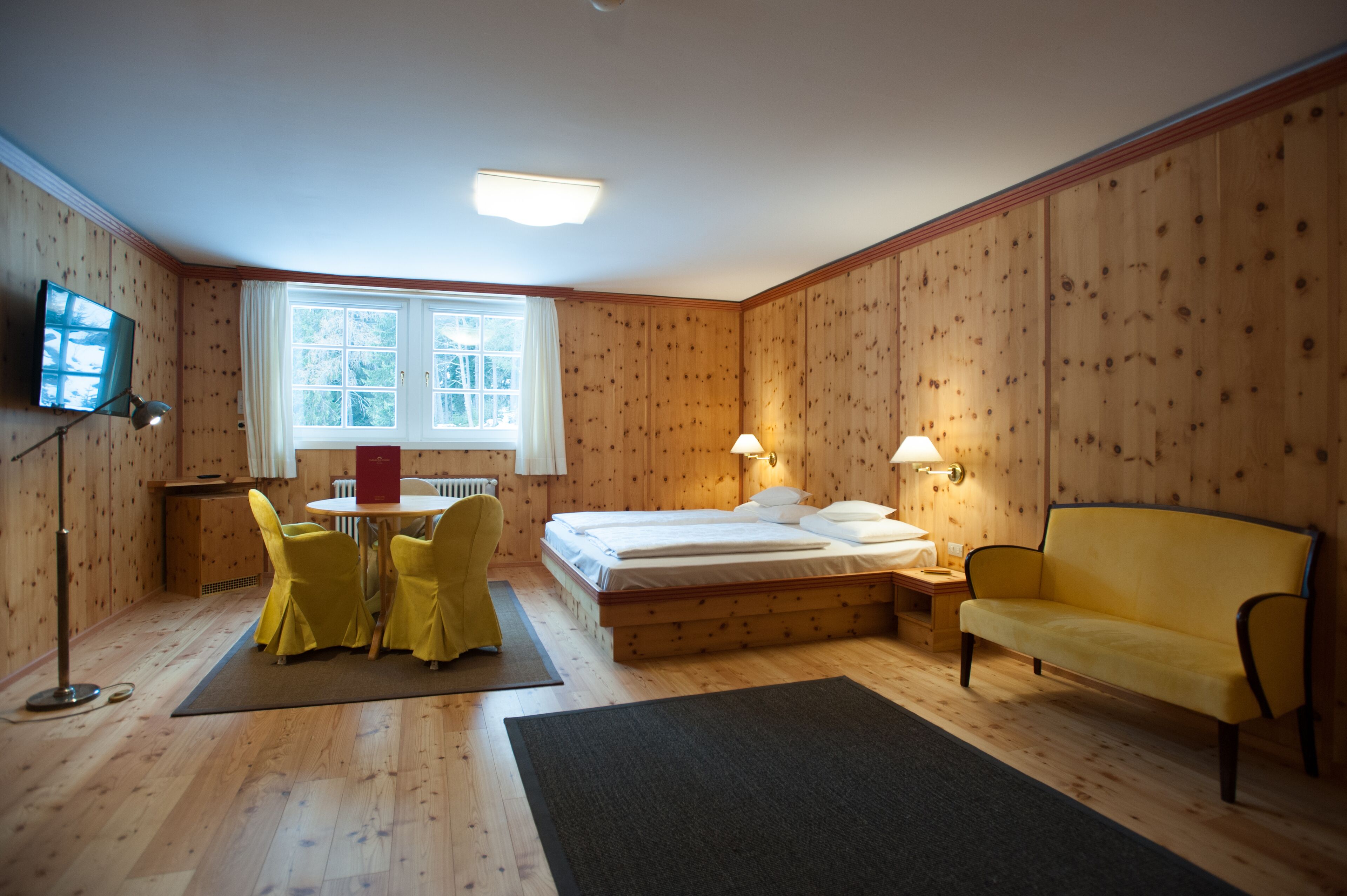 A room with a bed, a yellow sofa, a dining table with yellow chairs, a TV, and a window with curtains.