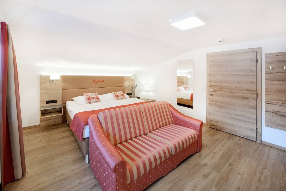 A hotel room with a bed, a striped sofa, a wooden wardrobe, and a mirror.