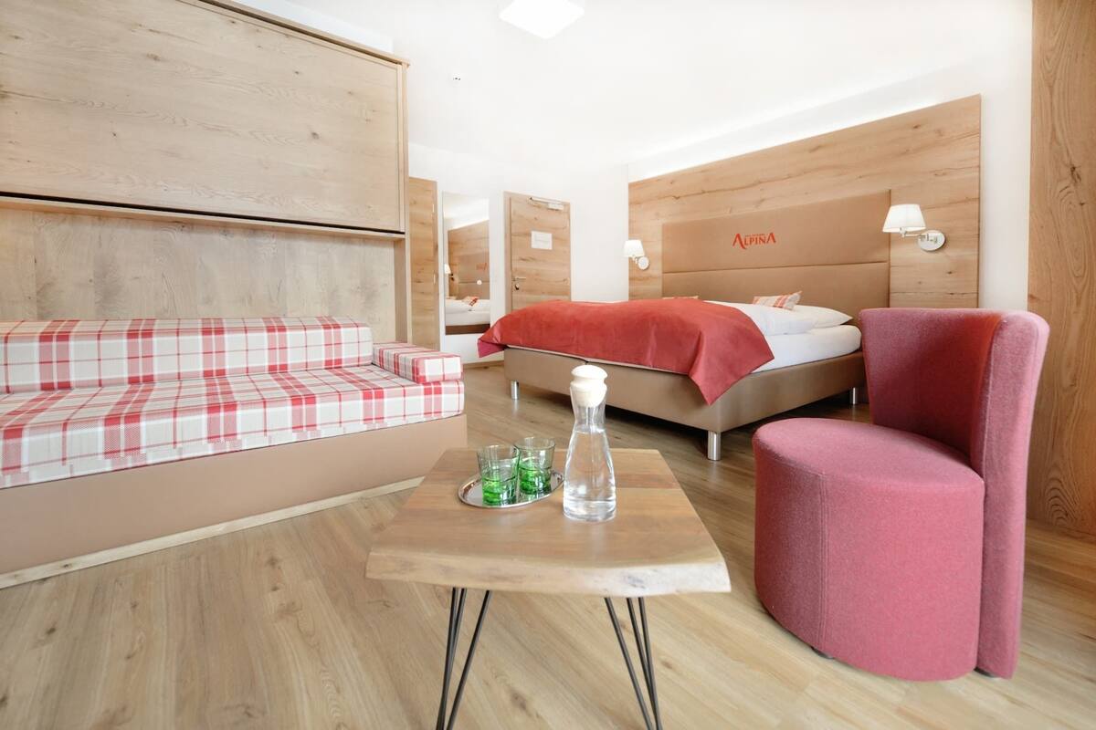 A modern hotel room with a wooden headboard, a red and white checkered sofa, a single bed with a red blanket, a pink armchair, and a small wooden table with a glass pitcher and glasses.