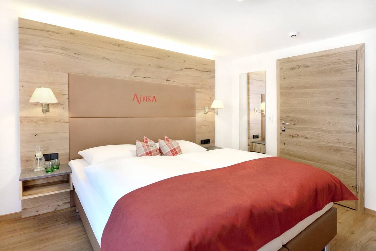 A hotel room with a bed, two bedside tables, a nightstand, a red throw blanket, and a wooden headboard with the brand name 'Alpina'.