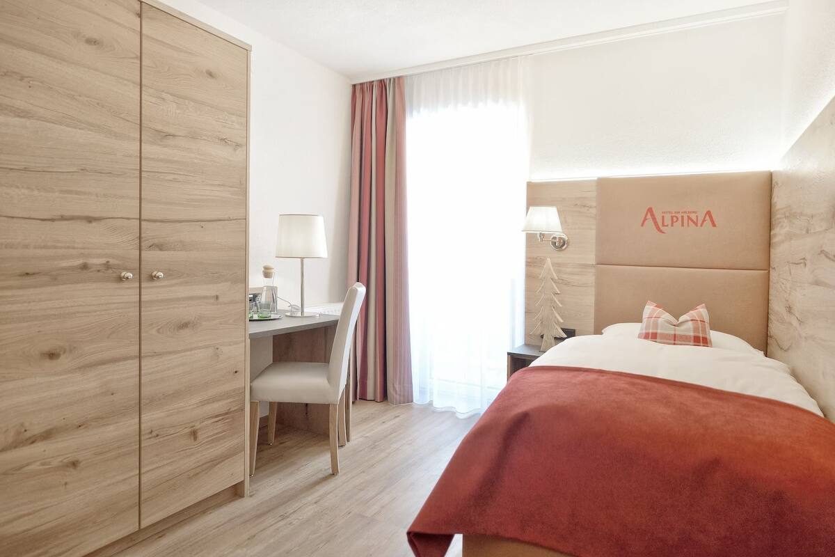 A hotel room with a bed, a desk with a chair, a lamp, and a window with curtains.
