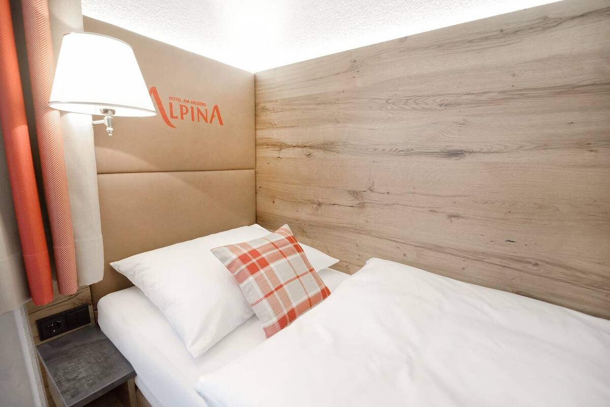 A hotel room with a wooden headboard, a bed with white bedding, a checkered throw pillow, and a lamp on a bedside table.