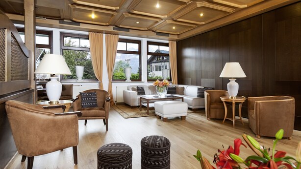 Una sala de estar moderna con un sofá, sillones, otomanas, una mesa de centro y lámparas. Amplios ventanales con cortinas ofrecen luz natural y vistas al exterior.