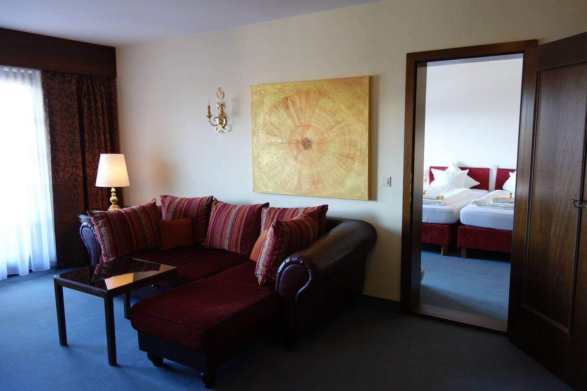 A hotel room with a dark red sofa, a small table with a lamp, a large abstract painting on the wall, and a doorway leading to a bedroom with two beds.