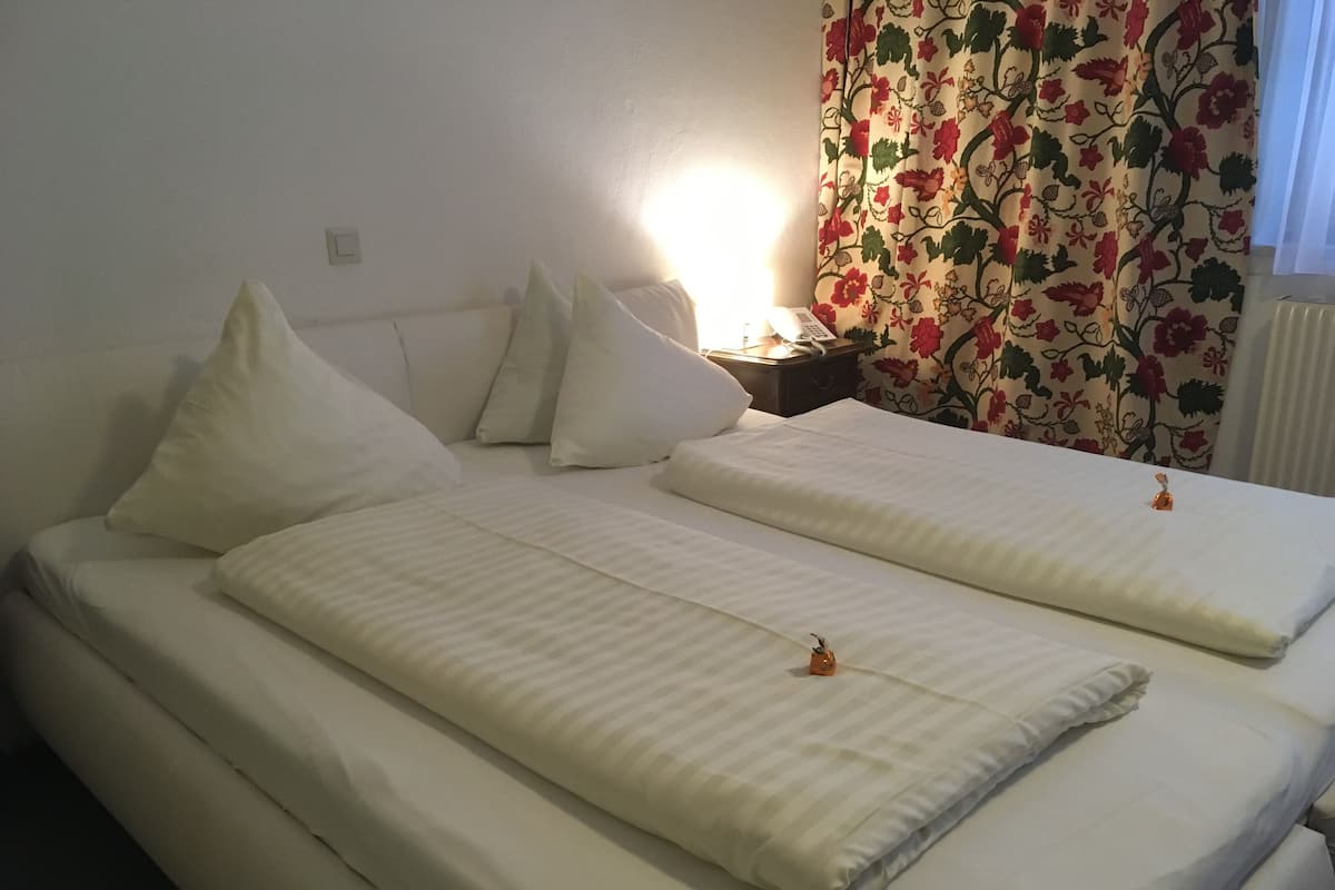 A double bed with white bedding and pillows, a bedside table with a lamp, and a floral curtain.