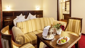 Habitación de hotel con cama, sofá, mesa de comedor con una bandeja de frutas, cubitera y un jarrón con flores.