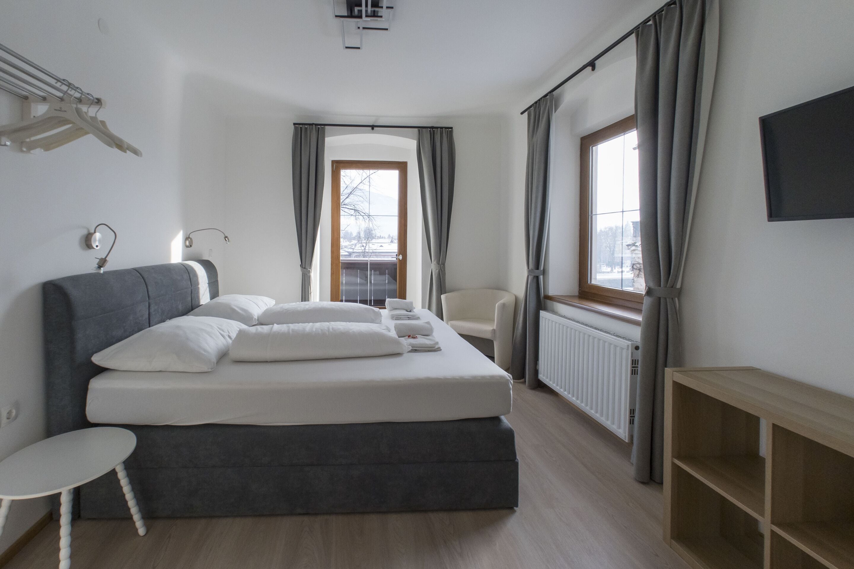 Un dormitorio moderno con una cama grande, una mesita, una silla, un televisor y una ventana con cortinas.