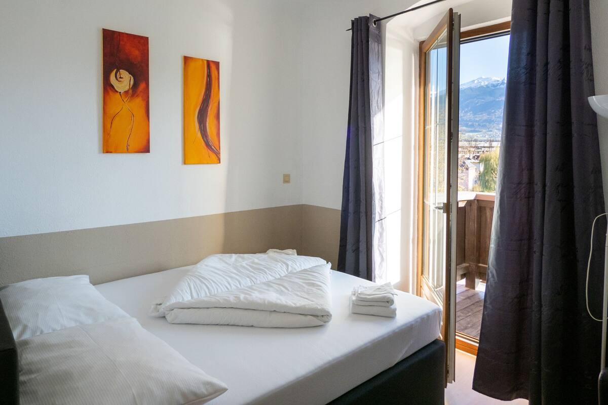 A hotel room with a bed, two paintings on the wall, and a view of the mountains through the window.