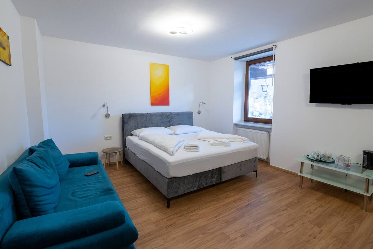 A modern bedroom with a grey bed, a teal sofa, a small side table, a glass shelf, and a TV on the wall.
