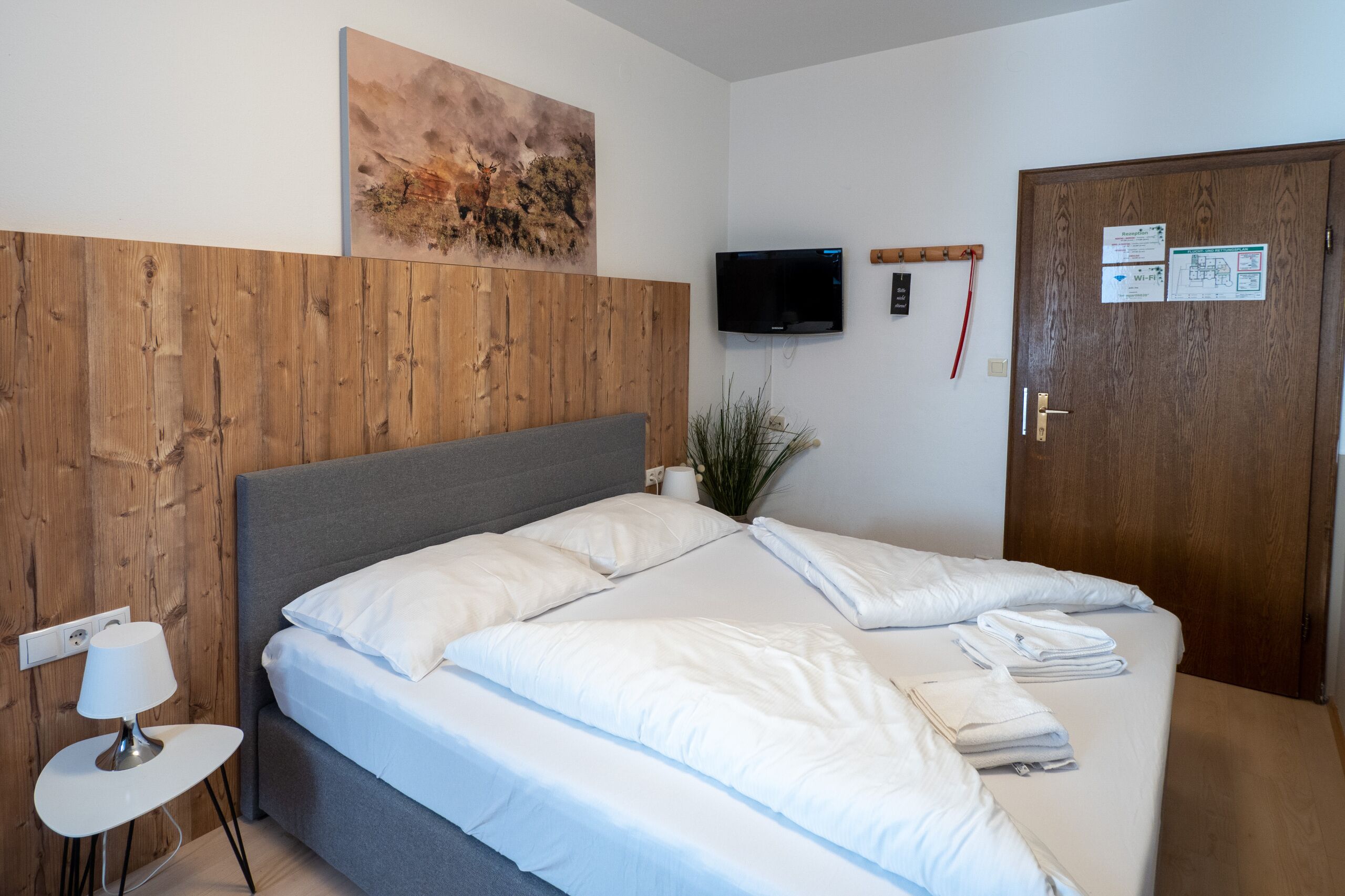 Quarto com cabeceira de madeira, televisão de tela plana na parede, porta com avisos e mesa de cabeceira com abajur.