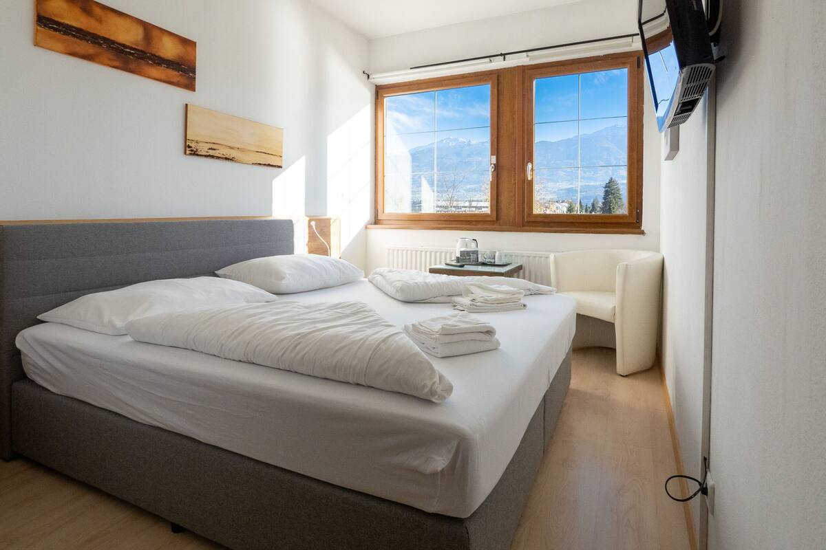 A bedroom with a large bed, a bedside table with a lamp, a window with a mountain view, and a wall-mounted TV.