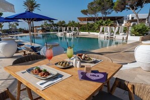 Zona de comedor junto a la piscina con una mesa para dos, piscina, sillones de descanso y sombrillas.