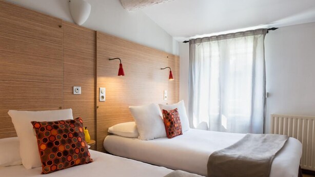 Une chambre d’hôtel avec deux lits, des lambris en bois aux murs, une fenêtre avec des rideaux légers et deux lampes suspendues rouges.