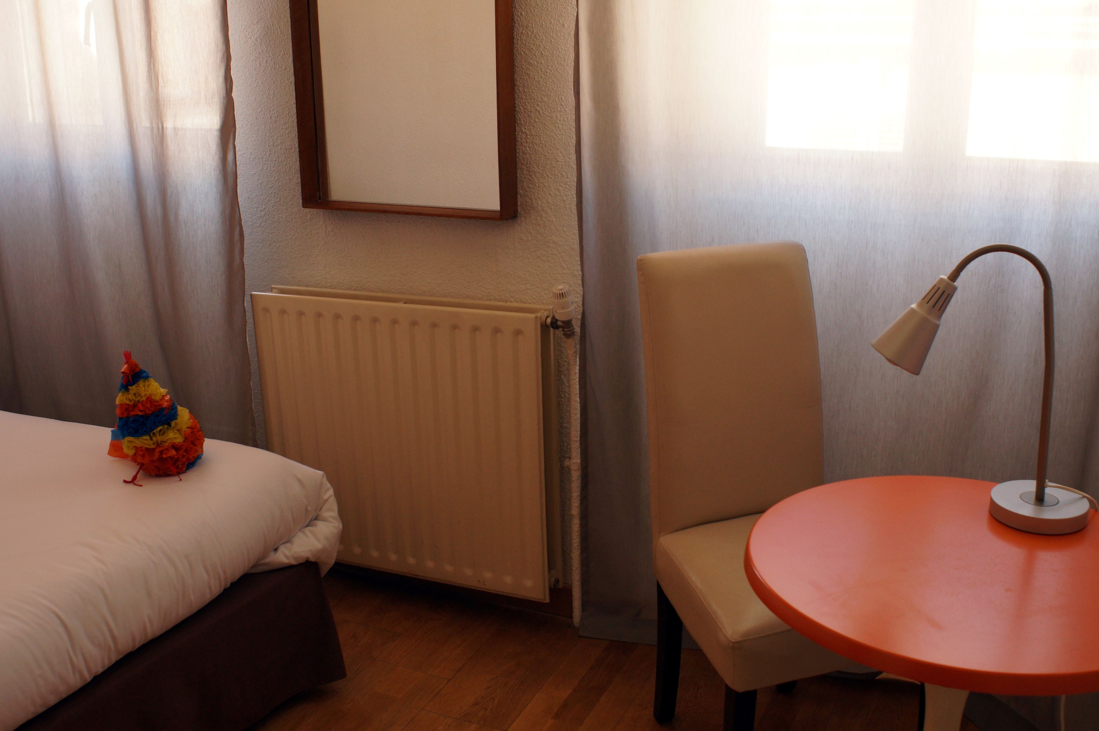 A hotel room with a bed, a chair, a small table, a radiator, and a window with curtains.
