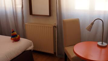 A hotel room with a bed, a chair, a small table, a radiator, and a window with curtains.