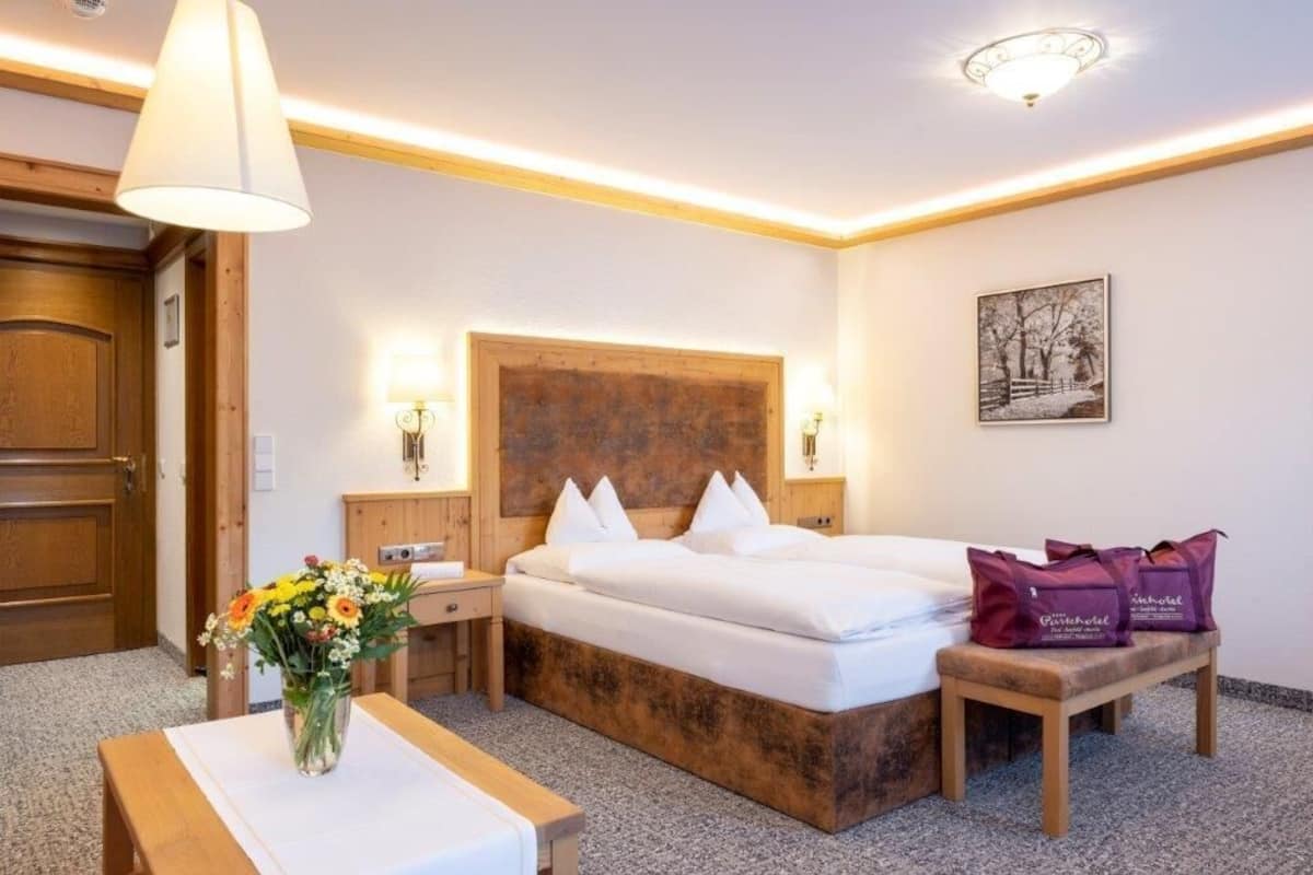 A hotel room with a wooden headboard, a bed with white linens, a bedside table with a vase of flowers, and a bench with purple bags.