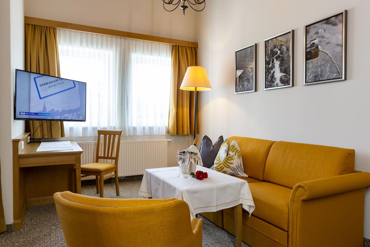 A hotel room with a desk, a computer, a sofa, a table with a lamp, and three framed pictures on the wall.