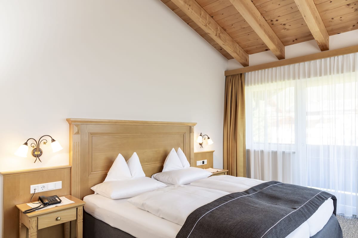 A hotel room with a wooden headboard, a bed with white linens, a bedside table with a lamp, and a window with sheer curtains.