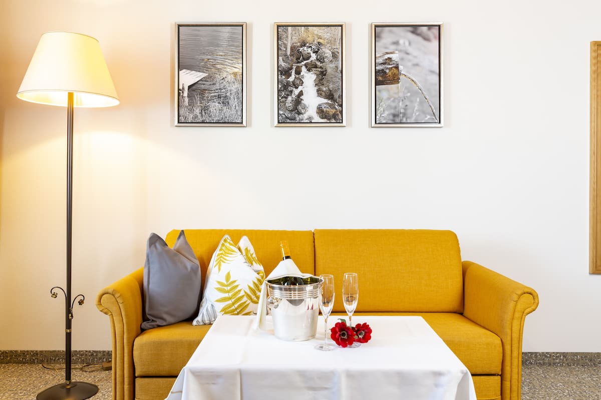 A room with a yellow sofa, a table with a bottle, glasses, and flowers, and three framed pictures on the wall.