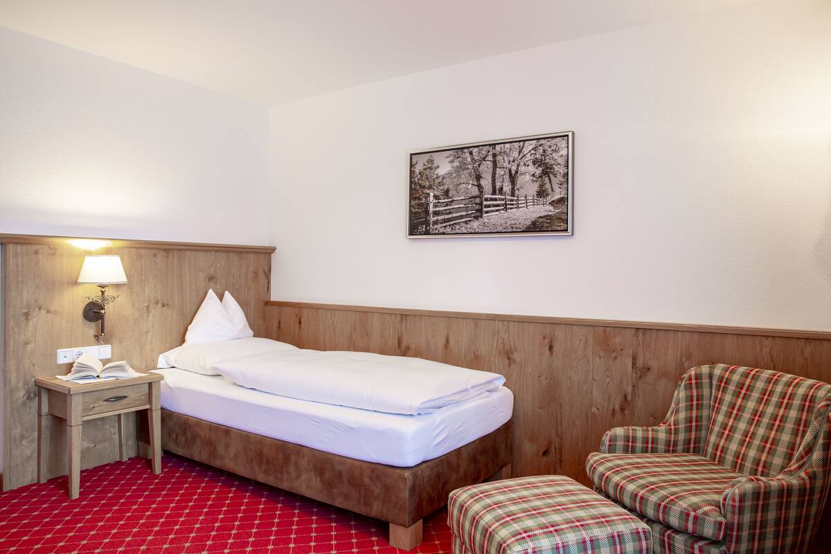 A hotel room with a wooden headboard, a single bed, a nightstand with a lamp, a plaid armchair, and a framed picture on the wall.