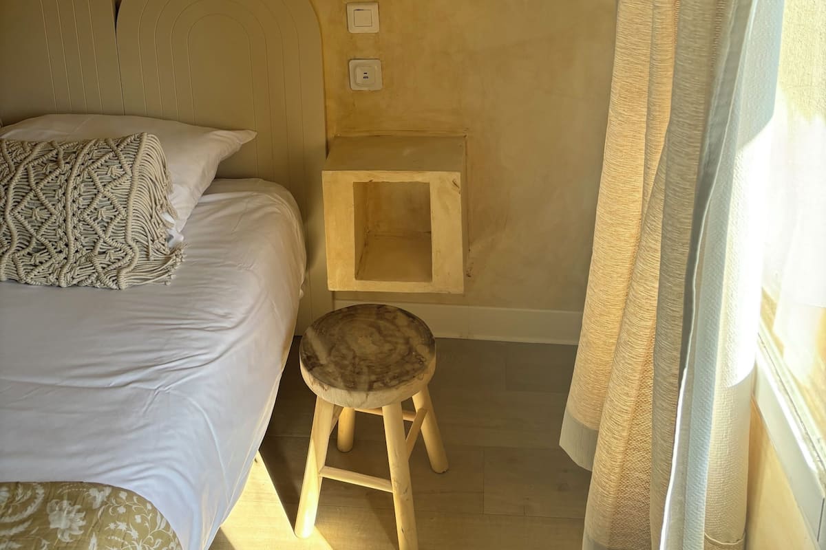 A bedroom with a bed, a small stool, a wall-mounted light, and a power outlet.
