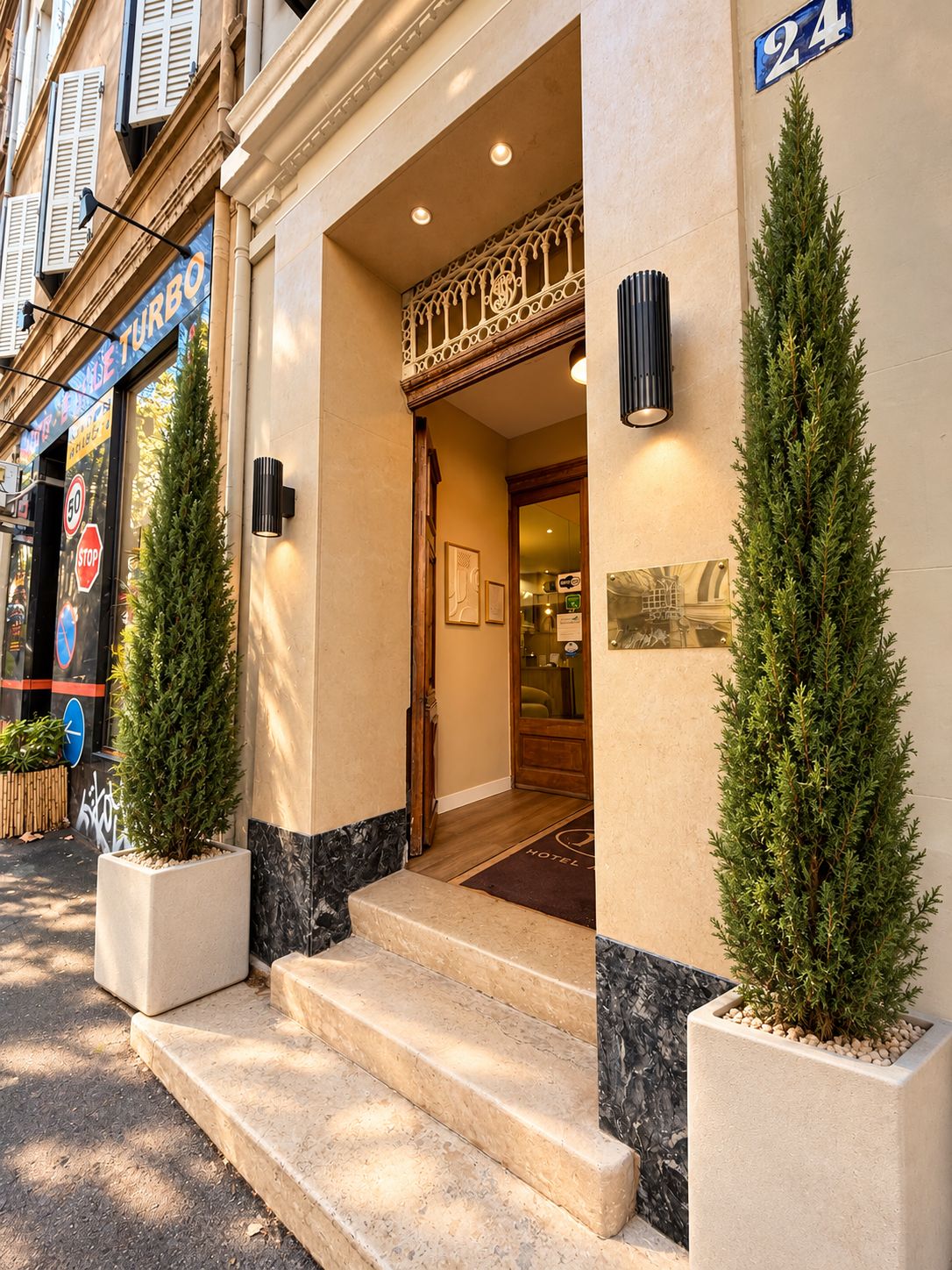 Entrada de hotel con puertas dobles, unos pocos escalones y dos árboles de hoja perenne en macetas.