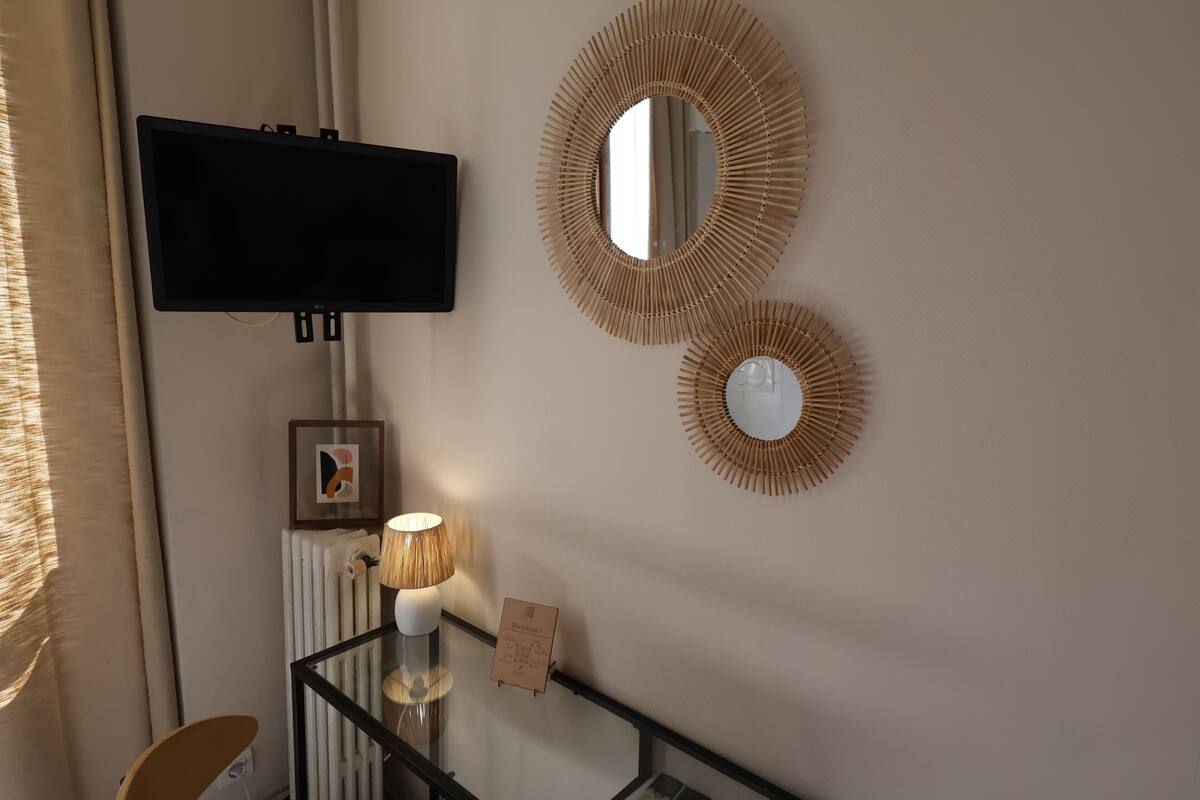 A room with a mounted television, two wall-mounted mirrors, a framed picture, a lamp on a glass table, and a radiator.