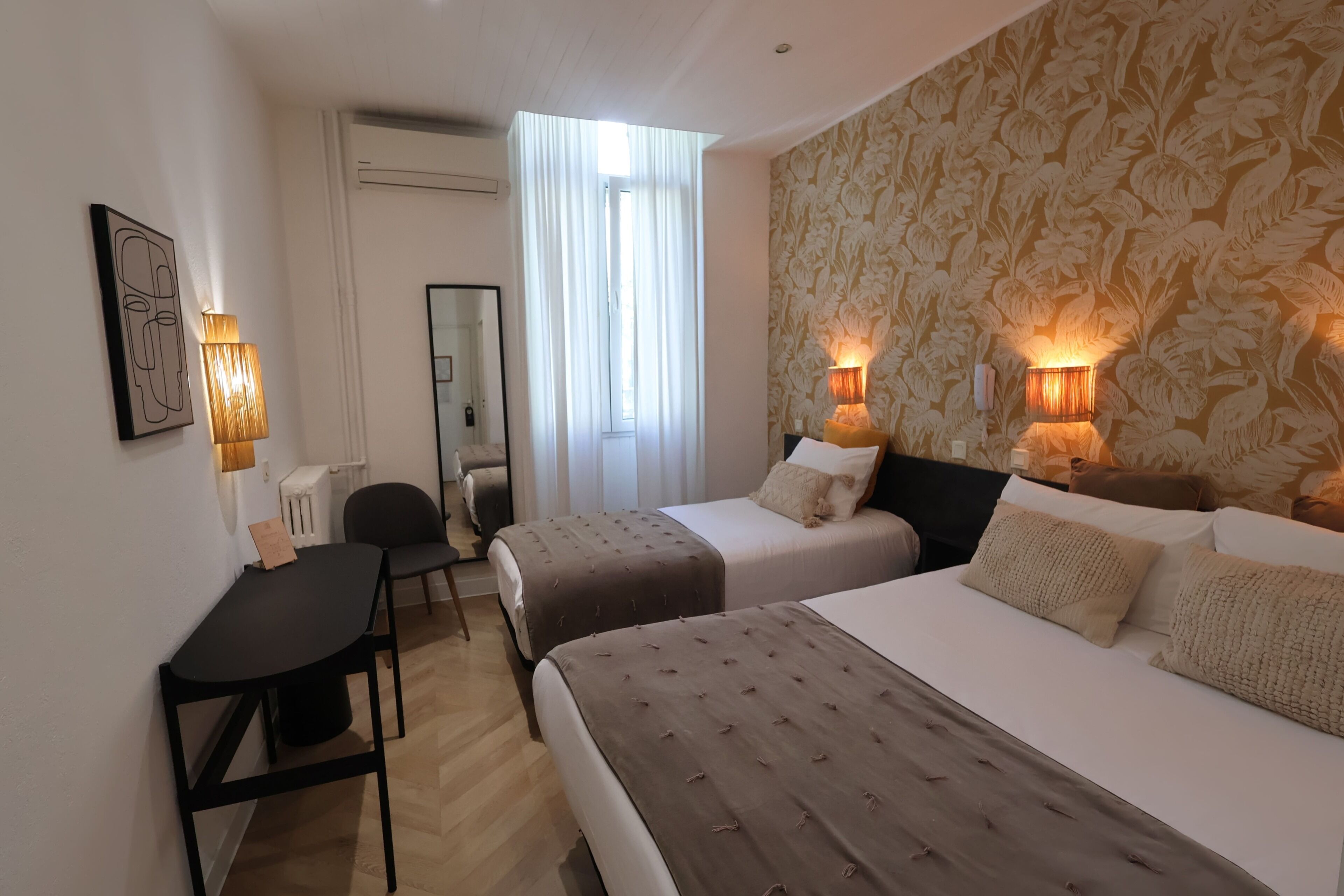 A hotel room with two beds, a desk, a chair, a mirror, and wall-mounted lamps.