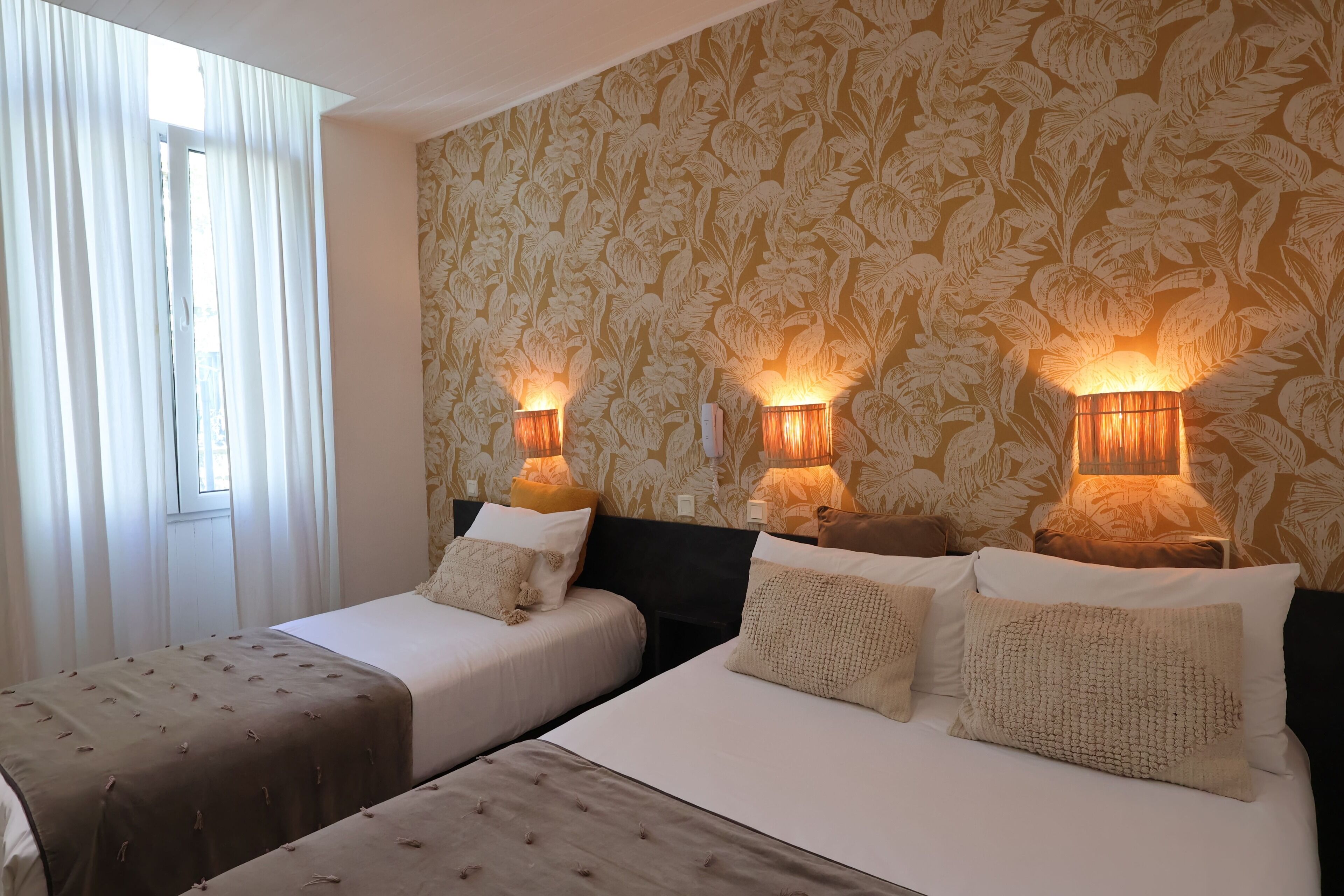 Habitación de hotel con dos camas, una ventana con cortinas, papel tapiz floral y lámparas fijadas a la pared.