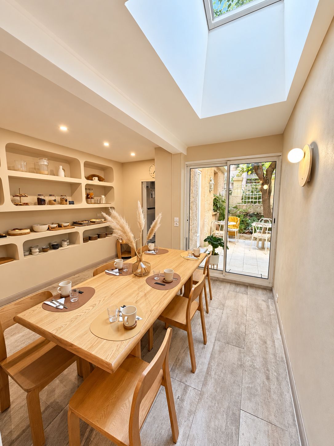 A dining area with a long wooden table, chairs, and a glass-top table. There are shelves with kitchenware and a view of the outside through a glass door.
