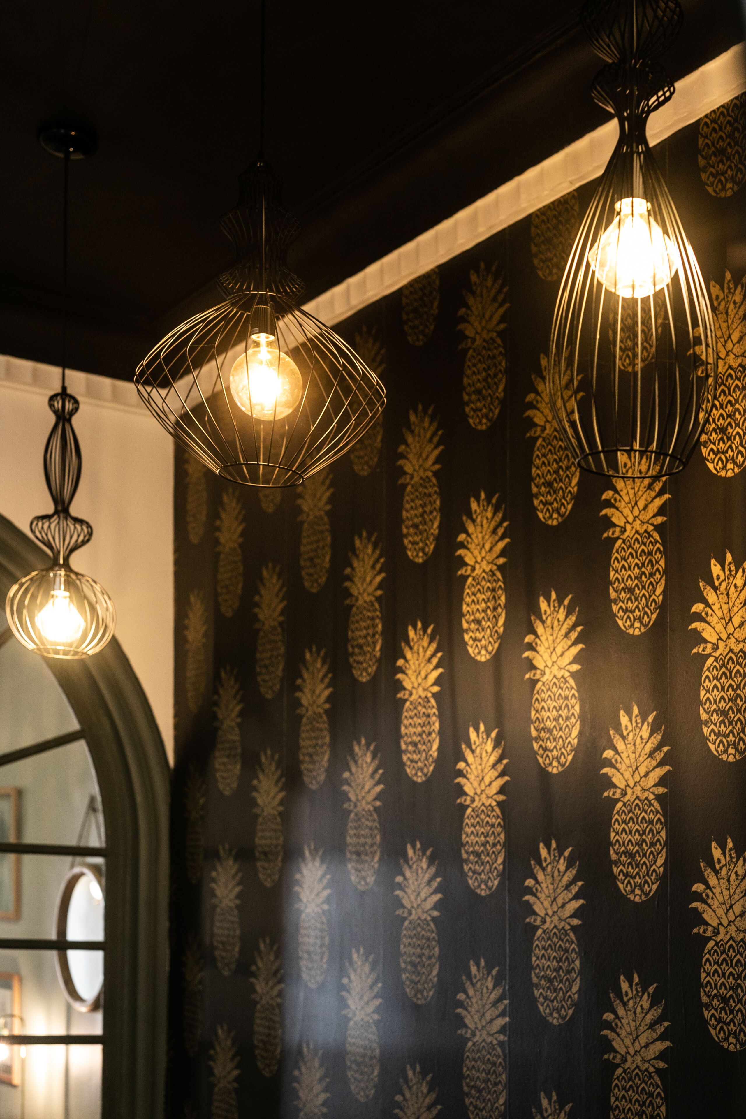 A room with pineapple-patterned wallpaper, hanging wireframe pendant lights, and a visible doorway leading to another area.
