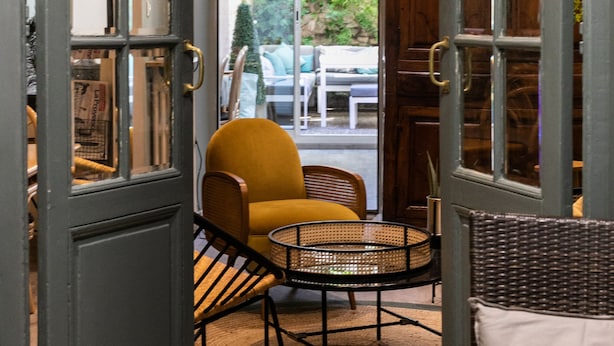 An interior with a large open doorway leading to a seating area with a yellow chair and a wicker table. - Azur (Marseille)