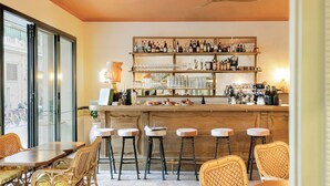 Zona de bar con estanterías de madera llenas de botellas, vasos y utensilios. En primer plano, sillas de mimbre y una mesa.