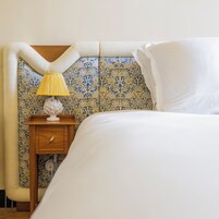 A neatly made bed with white linens, a small wooden nightstand with a lamp, and a decorative wall with a floral pattern.