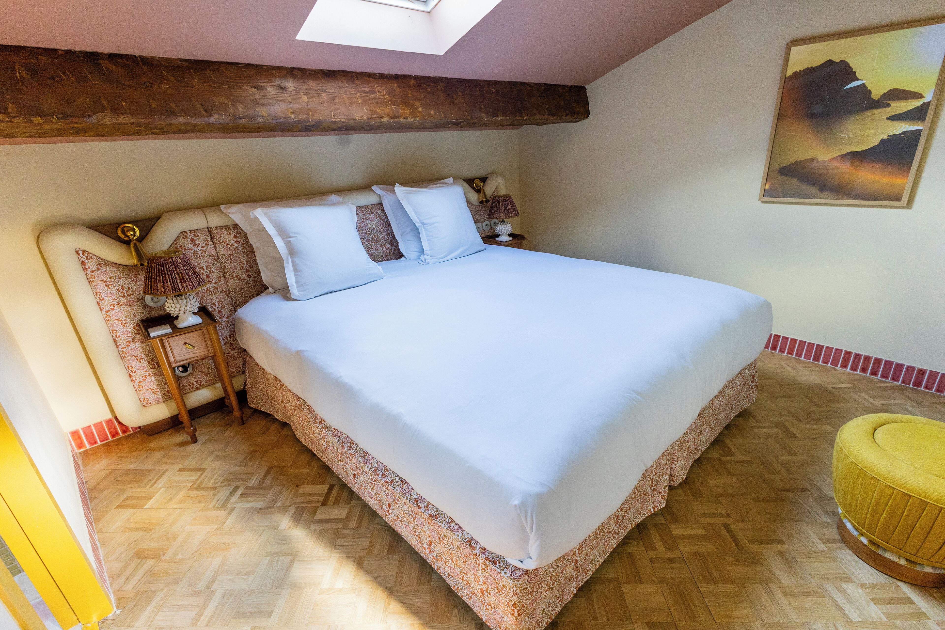 Un dormitorio con una cama grande, suelo de madera, un tragaluz, un cuadro y un pequeño taburete amarillo.
