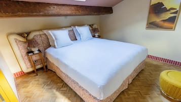 Un dormitorio con una cama grande, suelo de madera, un tragaluz, un cuadro y un pequeño taburete amarillo.