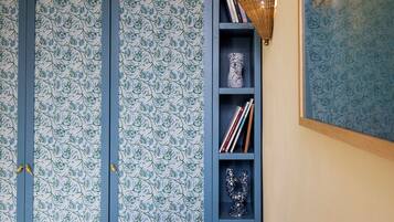 Een kamer met behang met een patroon, een boekenplank met boeken en decoratieve objecten, en een wandlamp.