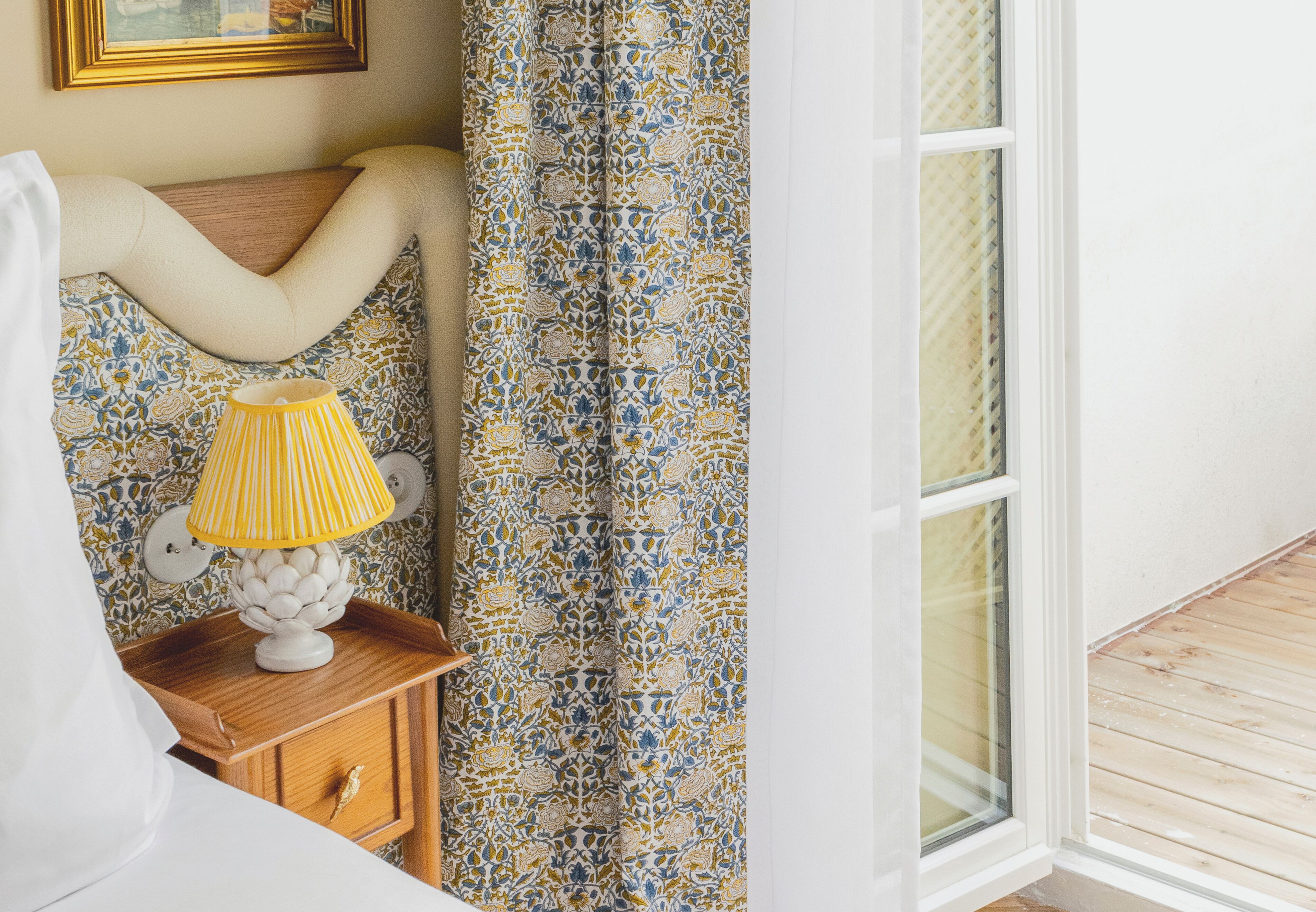 Un dormitorio con mesita de noche de madera, lámpara, cortinas estampadas y una ventana.