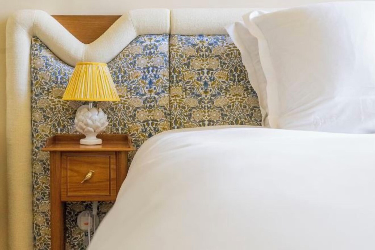Una cama bien hecha con una colcha blanca y almohadas, una mesita de noche de madera con una lámpara y un cabecero estampado.