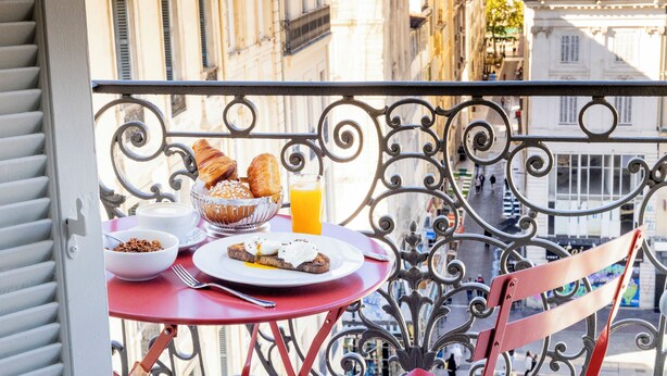 Un balcón con una mesa lista para el desayuno, que incluye croissants, un tazón de cereal y un vaso de jugo de naranja.