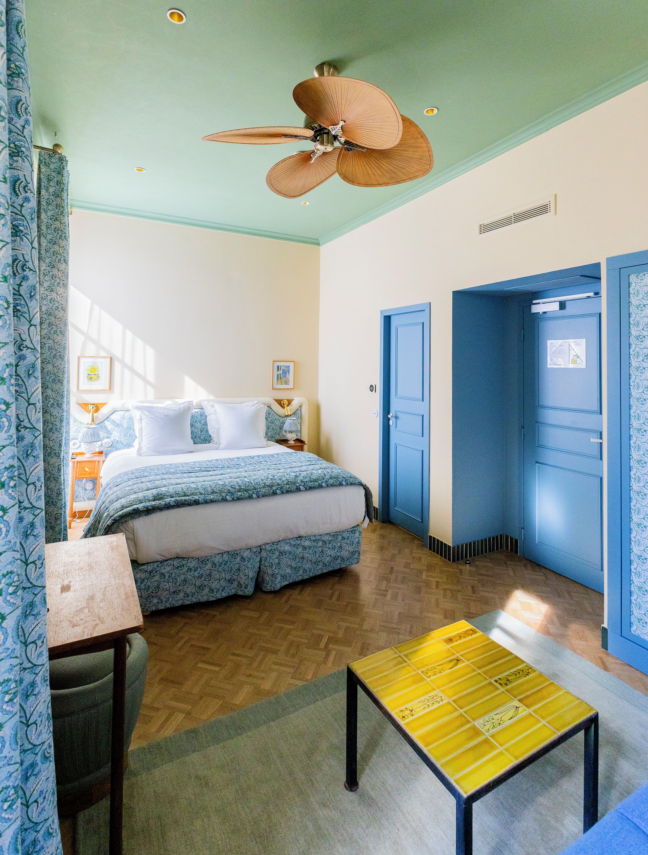 A bedroom with a bed, a desk, a chair, a ceiling fan, and two doors.