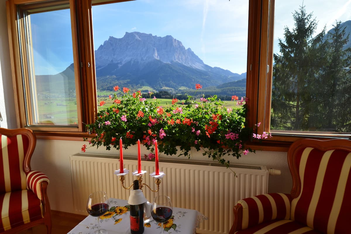 A room with a table set for two, a window with a view of mountains, and a potted plant with flowers.
