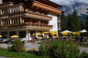 A hotel with outdoor seating and yellow umbrellas, surrounded by flowers and mountains in the background. - Sporthotel Zugspitze (Lermoos)