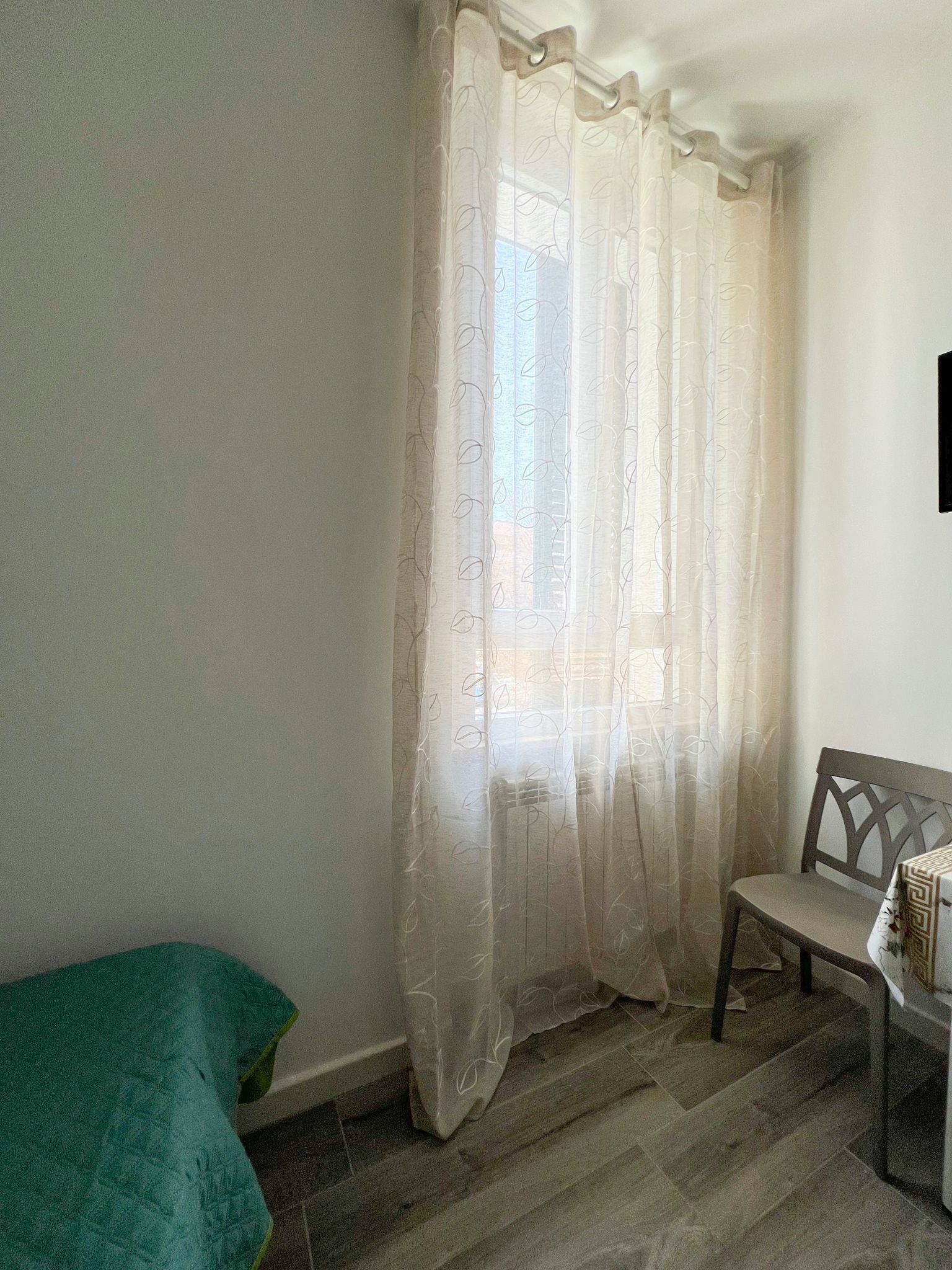 A room with a window covered by sheer curtains, a wooden chair, and a green blanket on the floor.