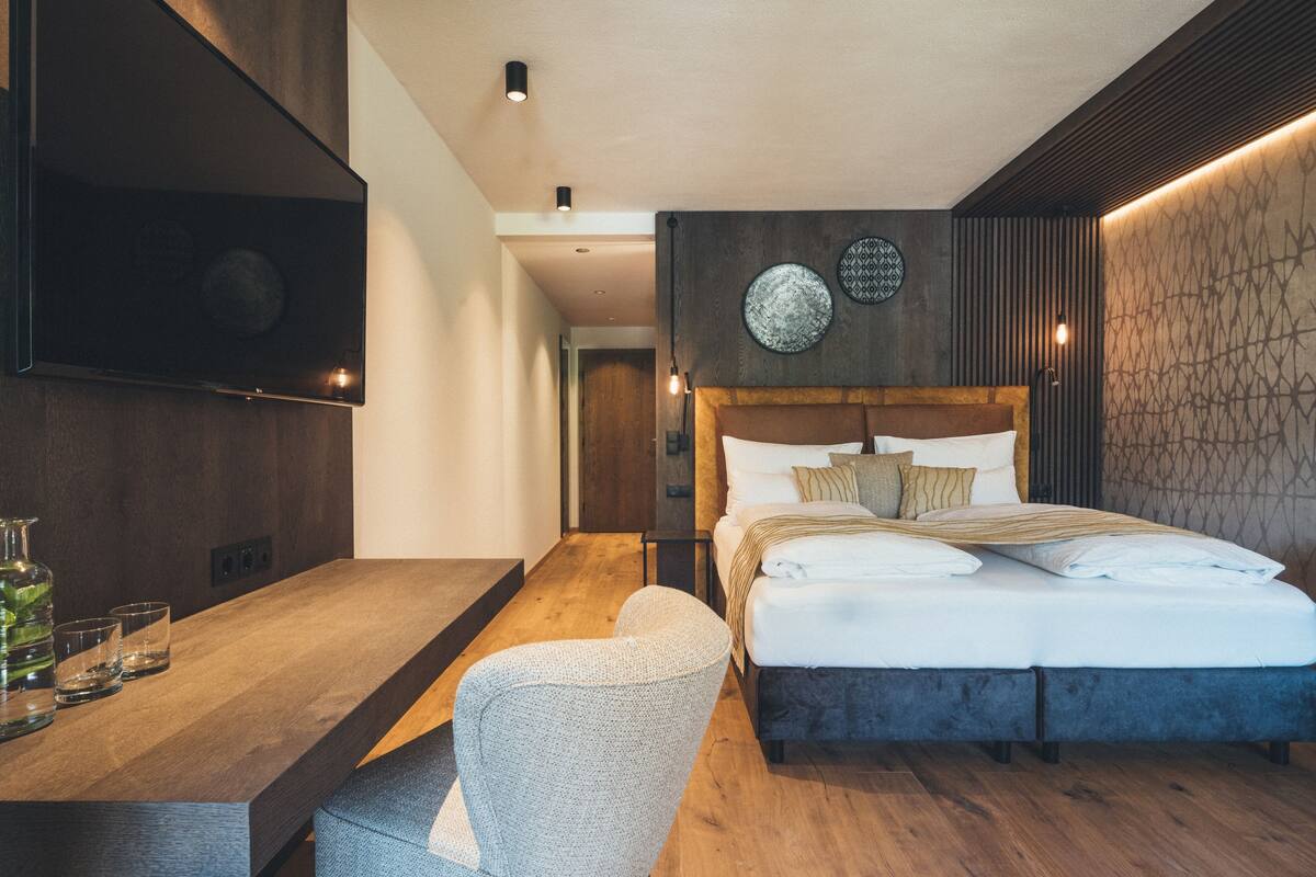 Una habitación de hotel moderna con suelo de madera, una cama grande con muchas almohadas, una mesa de comedor con vasos y un televisor montado en la pared.