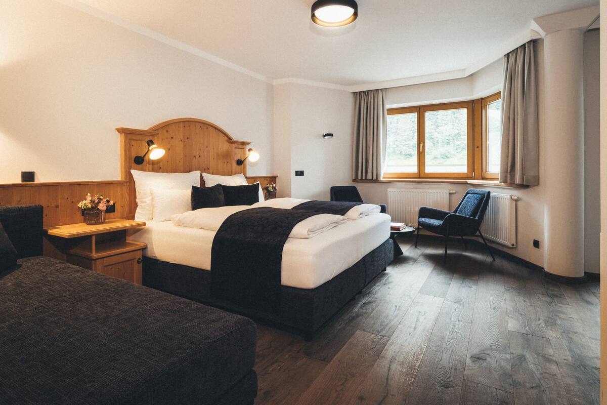 Una habitación de hotel con una cama grande, una mesita de noche con lámpara, una silla y una ventana con cortinas.