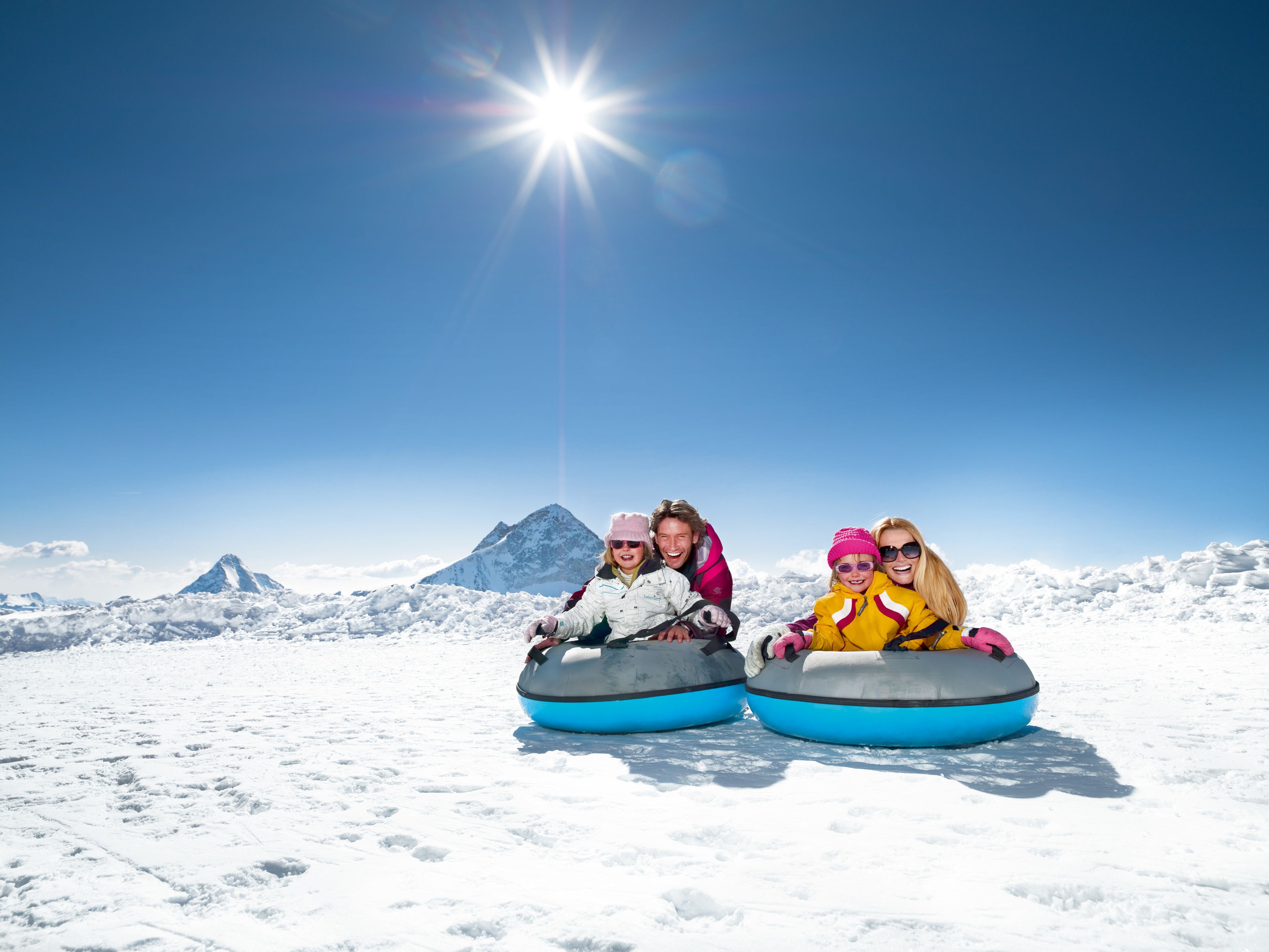 Three people on snow tubes on a snowy mountain with a clear sky and a sun shining.