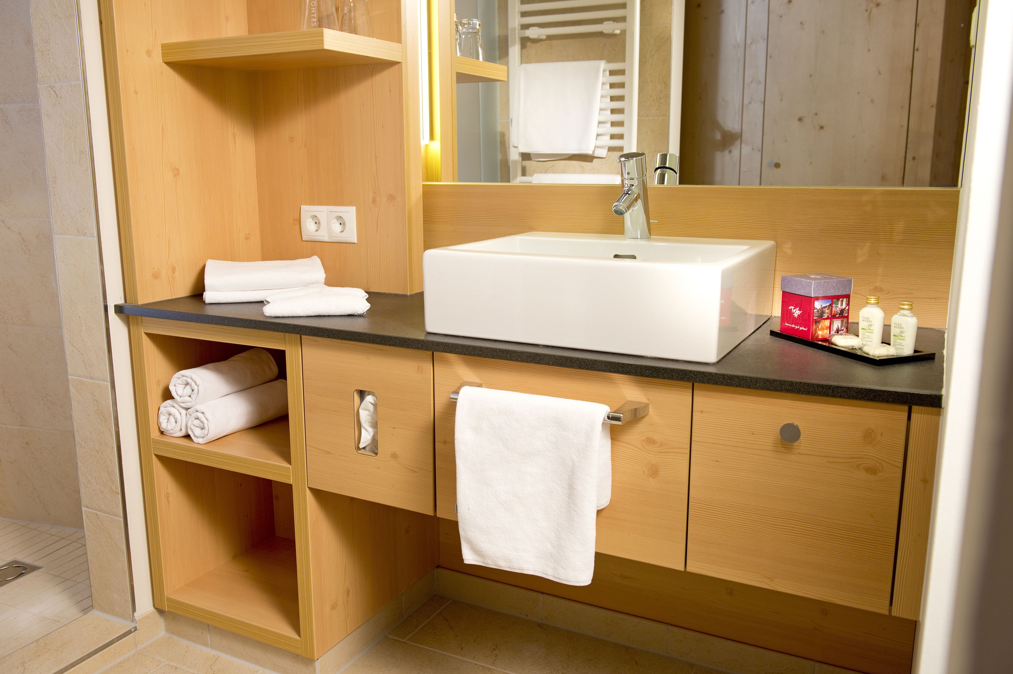 A modern bathroom with a wooden vanity, a white sink, and a mirror. There are towels and toiletries on the counter.